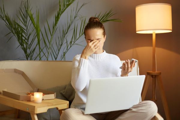 Mulher de meia idade, sentada no sofá, com um notebook no colo e o celular em uma das mãos, esfrega os olhos. Sinal claro da da Síndrome do olho seco.