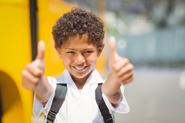 Garotinho negro sorridente, fazendo positivo com os polegares em razão da volta às aulas.