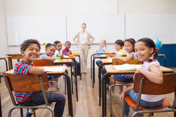 Sala de aula na qual a professora se encontra frente ao quadro branco e cujos alunos, sorridentes, olham para trás para fazer uma foto de turma.
