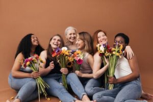 Seis mulheres de diferentes idades, peles e etnias, posam sorridentes segurando ramos de flores coloridas.