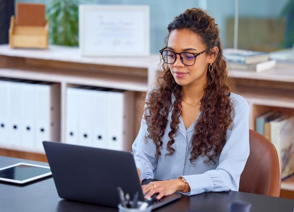 Mulher jovem, negra e de cabelos cacheados, trabalha em seu laptop no escritório perfeitamente iluminado.