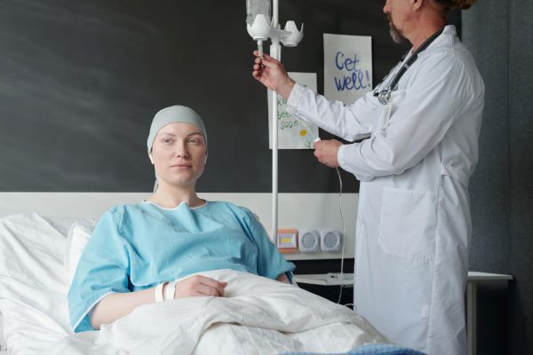 Mulher jovem, sentada em uma cama hospitalar, recebe o tratamento quimioterápico para o câncer de mama. Paciente veste um avental hospitalar azul e usa um lenço cinza claro.