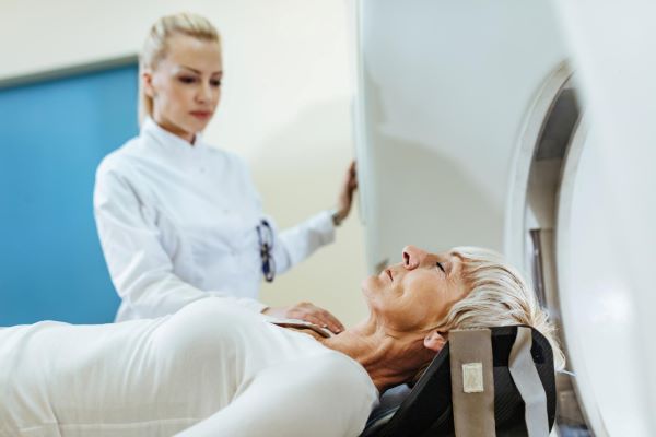 Mulher madura com cabelos curtos e grisalhos, deitada na esteira do aparelho de radioterapia, sendo preparada por uma técnica , que veste jaleco branco.