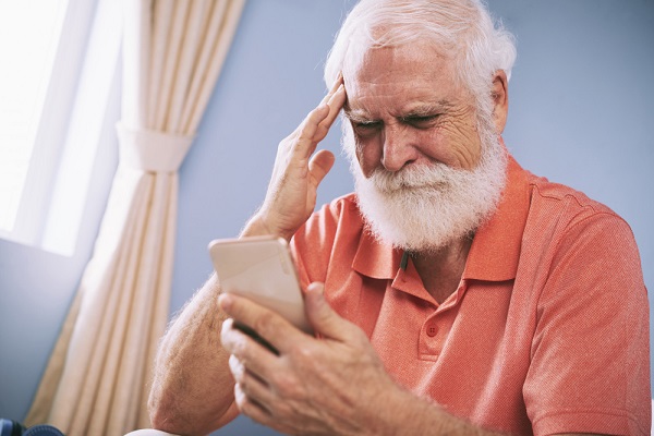 Idoso com cabelos e barba branca, vestindo uma camisa polo vermelha, coloca a mão na lateral da testa em sinal de esquecimento enquanto olha o celular.