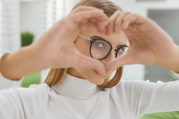 Linda jovem, loira com olhos claros, usando camiseta branca e óculos de grau, faz um coração com as mãos frente aos olhos. Conceito de saúde ocular.