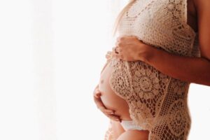 Gestante vestindo um vestido de crochê nude e segurando a barriga à mostra com as duas mãos.