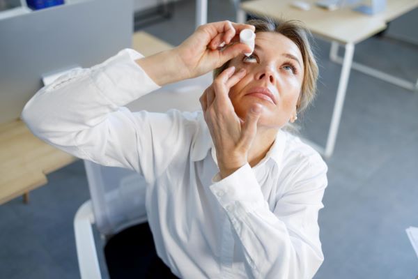 Linda mulher de meia idade, em seu ambiente de trabalho, pinga colírio lubrificante em seus olhos.