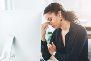 Empresária, frente ao computador em seu escritório, aperta a região dos olhos com a mão. Desconforto ocular em razão de estresse.