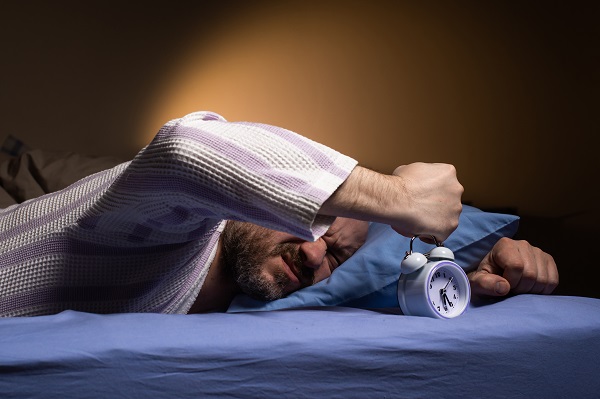 Homem maduro, deitado na cama, demostra irritação ao despertar querendo bater no despertador. 