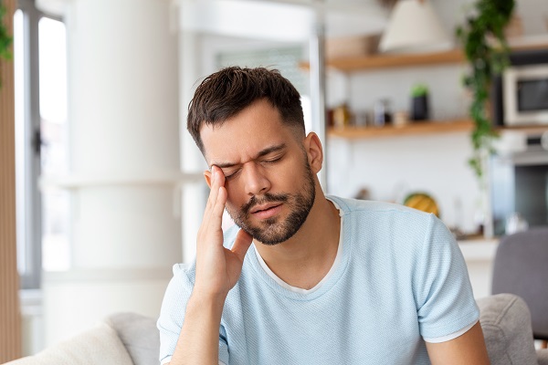 Homem bonito, com cabelos escuros e barba, colocando a mão nos olhos mostrando desconforto. Um dos sinais  do estresse é a enxaqueca.