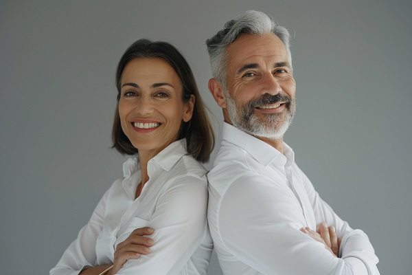 Casal maduro, um de costas para o outro, vestindo camisa branca, sorriem.