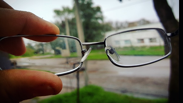 Pessoa segurando um óculos de grau através do qual se vê, de forma embaçada, uma rua com prédios e árvores. Um dos sintomas da miopia degenerativa é a visão turva, embaçada.
