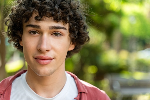 Close do rosto de um jovem com cabelos encaracolados, olhos escuros e uma pele livre de espinhas (acne).