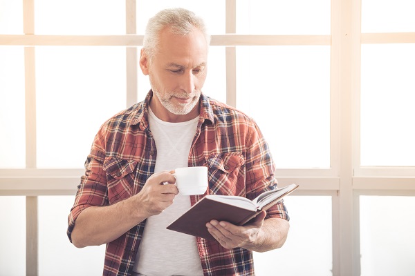Homem de meia idade, com cabelos grisalhos, vestindo t-shirt branca e camisa xadrez por cima, toma um café enquanto lê um livro sem a necessidade de óculos para perto.
Conceito de cirurgia no cristalino.