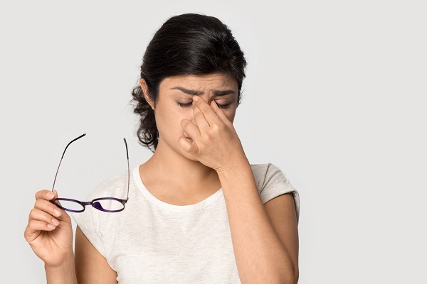 Garota jovem, com óculos pretos na mão direita, coça os olhos demonstrando sinais de desconforto ocular devido a alergia ocular.