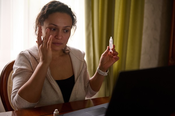 Mulher jovem, frente ao notebook, segura um frasco de colírio em uma das mãos após aplicá-lo nos olhos.