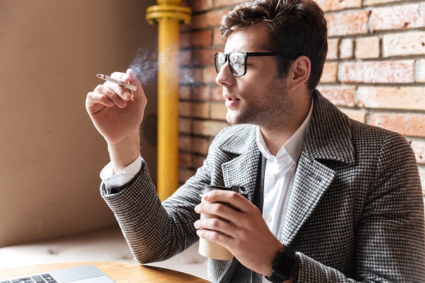 Jovem empresário, pensativo, bem vestido e usando óculos de grau, fuma um cigarro enquanto toma seu café.