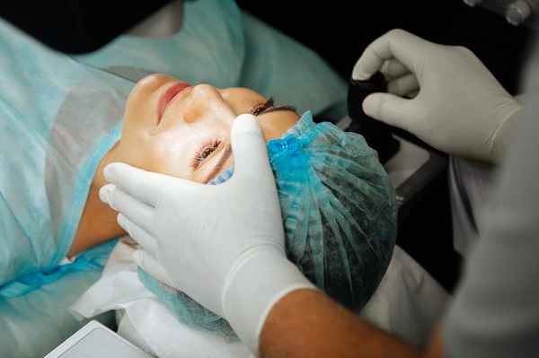 Paciente, na maca, sendo preparada para realizar cirurgia para ceratocone. Médico usando luvas brancas mexe em instrumentos acima dos olhos da mesma.