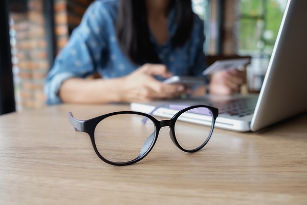 Close de um par de óculos de grau sobre uma mesa de trabalho na qual uma mulher está frente ao laptop.