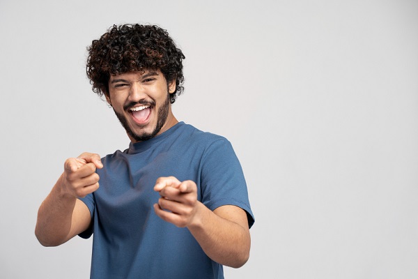 Jovem com cabelos cacheados e barba, vestindo camiseta azul, gesticula feliz após ter ganhado independência dos óculos e lentes de contato. 