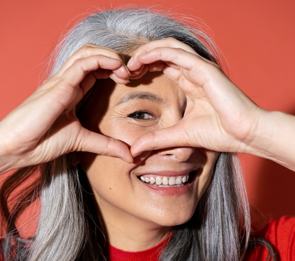 Mulher sorridente, com cabelos grisalhos e longos, faz um coração com as mãos sobre o olho direito.
