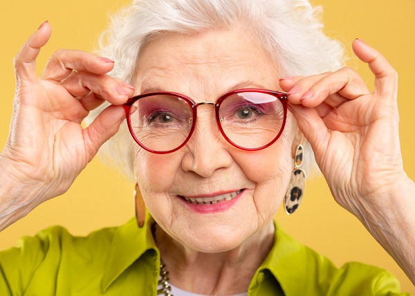 Bonita senhora idosa, vestindo roupas e acessórios modernos, com cabelos brancos e naturalmente maquiada, sorri feliz segurando um par de óculos com armação vermelha. 