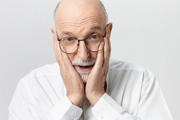 Idoso vestindo camisa branca e usando um par de óculos de grau pretos está com as duas mãos no rosto em sinal de preocupação.