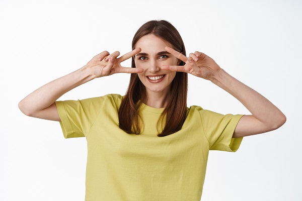 Linda jovem com cabelos longos e castanhos, vestindo uma camiseta amarela, destaca os olhos perfeitos com as duas mãos. 