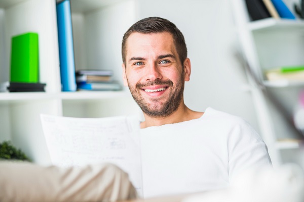 Homem jovem, bonito, com olhos claros, barba e cabelos escuros, vestindo camiseta branca, sorri satisfeito enquanto avalia documentos em seu escritório.