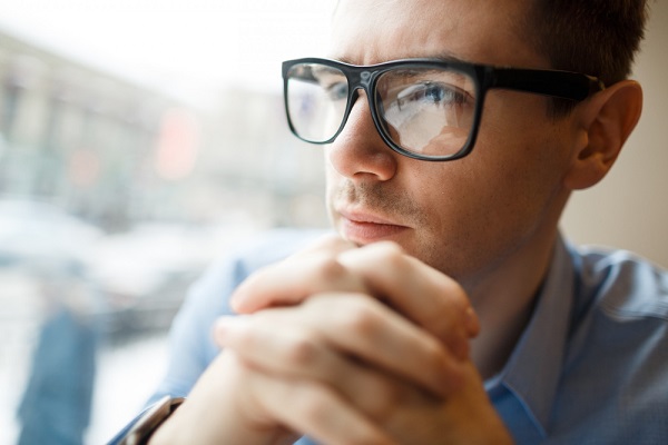 Jovem empresário, sério, observando a rua. Ele usa camisa azul e óculos de grau pretos.