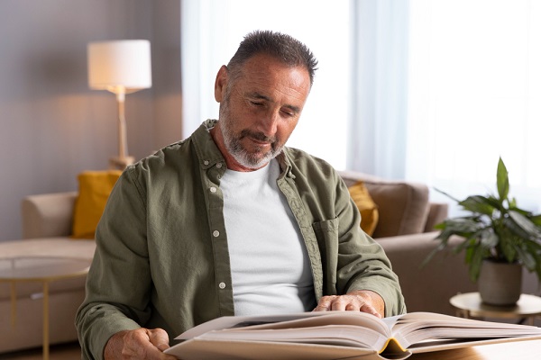 Homem de meia idade, grisalho com barba, vestindo camiseta branca e, sobre ela, uma camisa caqui, lê um livro sem o auxílio de óculos de grau.