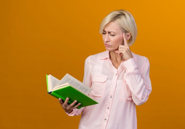 Mulher de meia idade, loira, vestindo camisa rosa, segura um livro de capa verde volumoso com o braço direito esticado, sugerindo vista cansada ou presbiopia.