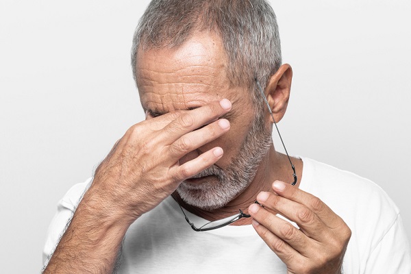 Homem idoso, com barba e cabelo grisalhos, esfrega os olhos com as mãos em sinal de desconforto. 
Um dos sintomas dos carcinomas é a sensação de desconforto ocular,