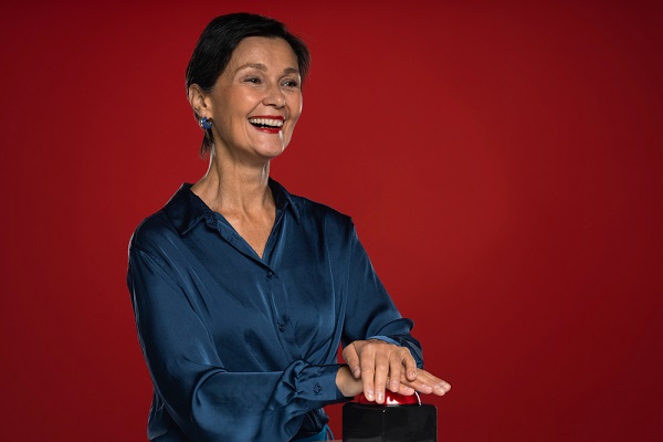 Mulher de meia idade, charmosa, com cabelos curtos , maquiada e vestindo uma camisa de seda azul, sorri feliz participando de um jogo.