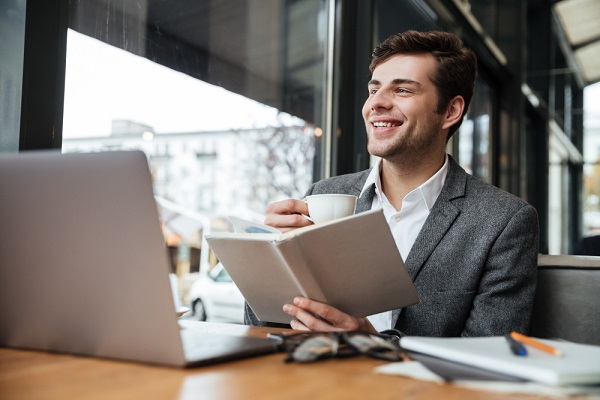 Jovem empresário, frente ao notebook, sorri enquanto bebe café e lê sua agenda. 
Um dos exercícios que evitam a fadiga ocular é afastar os olhos da tela por alguns instantes.